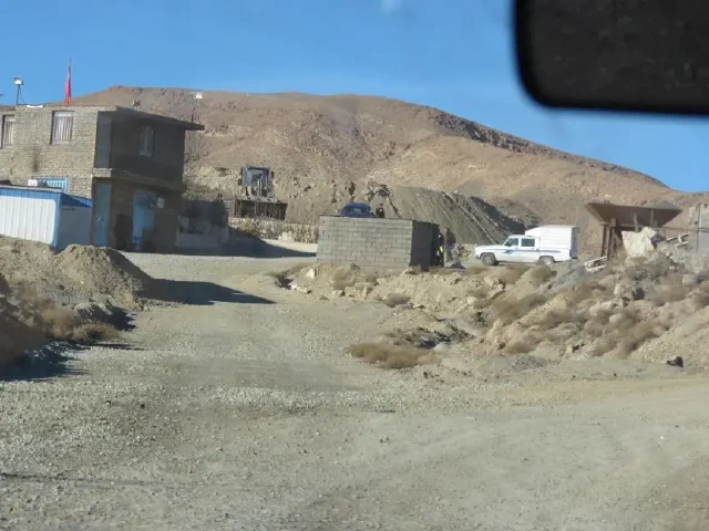 Buildings and a white van beside a dirt road below a bare hillside