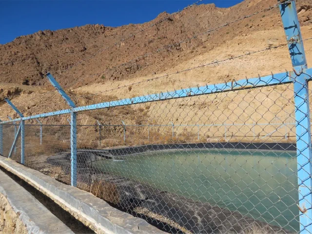 Water storage pool behind a blue chain-link fence with a rocky hillside beyond