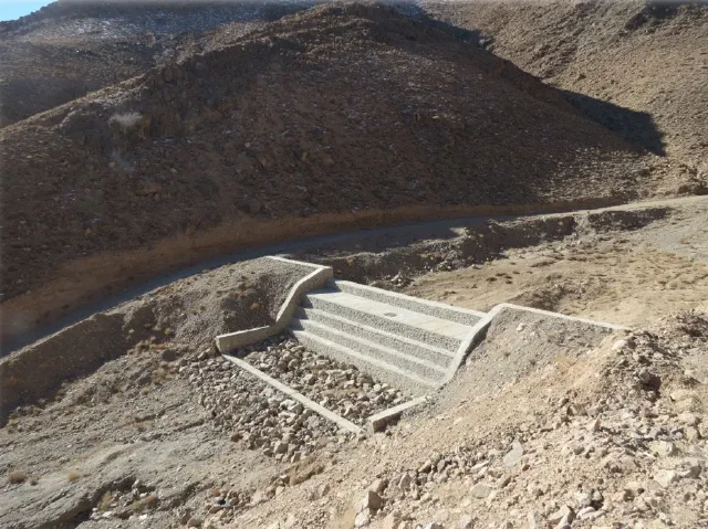 Stepped concrete barrier across a dry rocky channel beneath a hillside